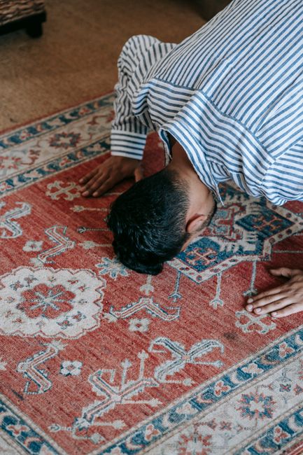 A person with dark hair wearing a blue and white striped shirt is kneeling on a richly patterned Persian-style carpet, pressing their forehead to the surface as part of a cleaning or sanitising process. The carpet features a red base with intricate floral and geometric motifs in shades of blue, cream, and light brown, and appears well-maintained and clean. The room has warm lighting, and the surrounding floor is visible with a section of brown carpet. Bow Carpet Cleaning offers professional deep cleaning services for carpets like this, ensuring hygiene and freshness in residential spaces. The image highlights surface cleaning and sanitisation techniques used to maintain the condition of domestic carpets.