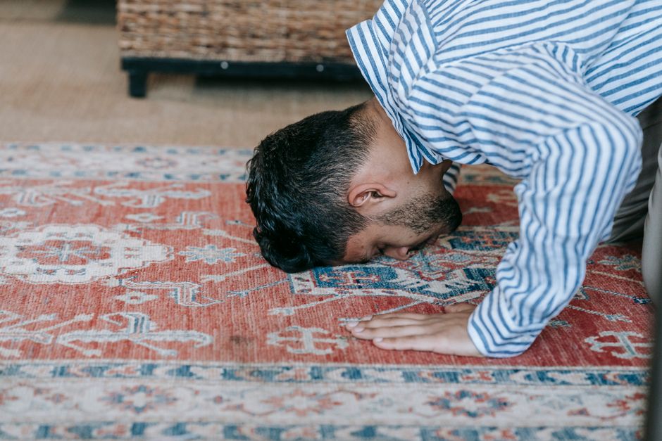 A man in a blue and white striped shirt is performing a deep cleaning or surface sanitisation of a patterned area rug in a well-lit room, using his hands and face to meticulously clean the intricate designs and fibers of the carpet. The rug features a traditional floral and geometric pattern with shades of red, blue, and cream. The background shows a wooden floor and part of a dark wooden piece of furniture. Bow Carpet Cleaning offers professional carpet cleaning services near Bow Road station, BOW, ensuring hygiene and freshness for domestic and commercial surfaces.