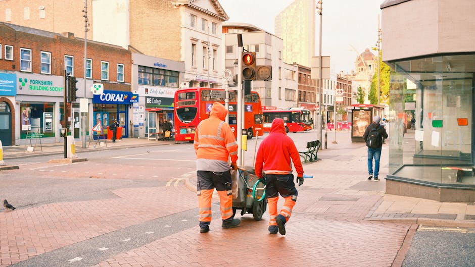 Two street cleaning workers in high-visibility orange and red uniforms are walking on a paved pedestrian area in Bromley by Bow, London. They are pushing a cleaning cart equipped with brushes and cleaning tools. The background shows a busy city street with commercial buildings, including shops and a bus stop, and a red double-decker bus approaching a traffic light. The scene is well-lit with natural daylight, and the area appears clean and well-maintained, reflecting professional cleaning services by Bow Carpet Cleaning as part of urban hygiene and surface maintenance for commercial environments.