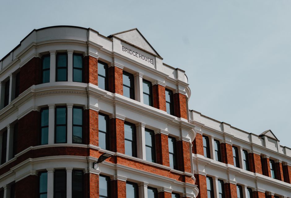 The image shows the upper floors of a historic red brick building with white decorative trim and large, evenly spaced windows. The building, labeled 'Bridge House,' displays characteristic Victorian architectural details with rounded corners and ornate window frames. The exterior surfaces are clean and well-maintained, with the brickwork appearing fresh and free of dirt or grime. The white trim contrasts sharply with the red brick, highlighting the detailed architectural features. Bright natural daylight illuminates the façade, emphasizing the building's structure and pristine appearance. This image showcases the importance of regular surface cleaning and maintenance for historic buildings, aligning with Bow Carpet Cleaning's expertise in comprehensive cleaning services for commercial properties. The overall scene reflects a tidy, well-preserved urban building, suggesting professional cleaning and upkeep contribute to its attractive appearance.
