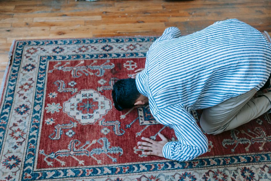 A person kneeling on a patterned red and cream rug in a room with wooden flooring, engaged in cleaning or prayer. The individual is wearing a blue and white striped shirt and beige trousers, with their head bowed and hands on the rug. The rug features intricate floral and geometric motifs, and the room is well-lit, emphasizing the cleanliness and detail of the surface. Bow Carpet Cleaning provides expertise in deep cleaning and sanitisation of domestic surfaces, ensuring hygiene and freshness in residential spaces, particularly around Bow Church, BOW.