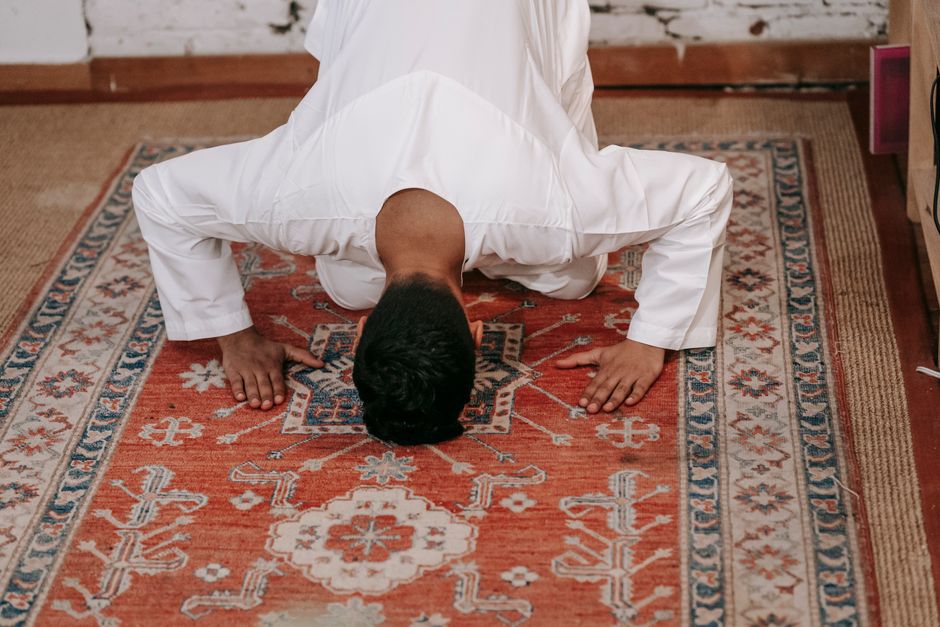 A person dressed in white, kneeling on a patterned red and beige oriental rug, performing a floor cleaning or deep cleaning process. The individual is positioned with their head touching the ground and their hands placed on the rug, showcasing a focused cleaning activity. The room features wooden floors surrounding the rug, with natural lighting illuminating the space. Visible in the background are part of the wooden wall and a piece of furniture, suggesting a cozy, home interior. This image demonstrates surface cleaning techniques suitable for maintaining cleanliness and hygiene, consistent with services provided by Bow Carpet Cleaning near Bow Church, BOW.