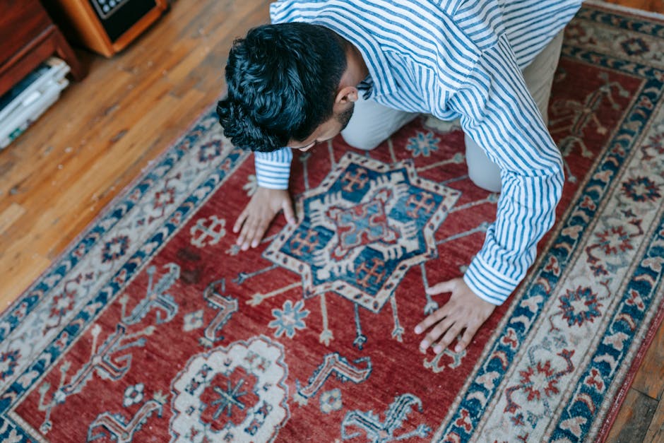 A person kneeling on a patterned red and cream rug in a room with wooden flooring, engaged in cleaning or prayer. The individual is wearing a blue and white striped shirt and beige trousers, with their head bowed and hands on the rug. The rug features intricate floral and geometric motifs, and the room is well-lit, emphasizing the cleanliness and detail of the surface. Bow Carpet Cleaning provides expertise in deep cleaning and sanitisation of domestic surfaces, ensuring hygiene and freshness in residential spaces, particularly around Bow Church, BOW.
