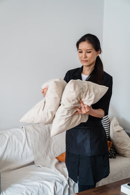 A woman with dark hair dressed in a black uniform is holding two beige cushions while standing in a bright living room. The room features a white sofa with several cushions, including a black and white patterned one, and a wooden table partially visible in the foreground. The background includes plain white walls and natural lighting, creating a clean and tidy environment that suggests professional domestic cleaning services. The scene emphasizes surface cleaning and maintaining hygiene in a residential setting, consistent with upholstery deep cleaning for E3 homes in Devons Road, Bow, provided by Bow Carpet Cleaning.